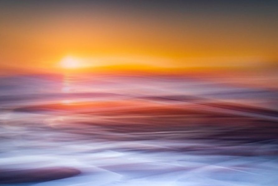 Abstract, blurred long-exposure photograph of the ocean at sunset, featuring vivid streaks of orange, red, and electric blue water movement.