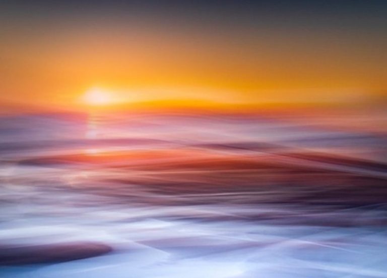 Abstract, blurred long-exposure photograph of the ocean at sunset, featuring vivid streaks of orange, red, and electric blue water movement.